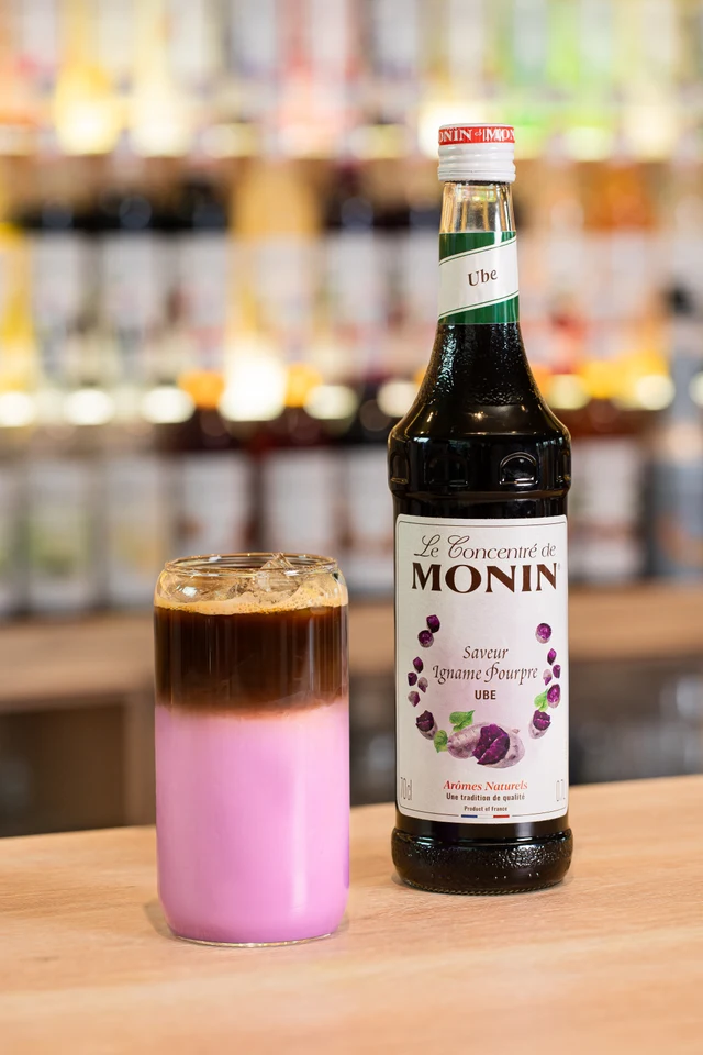 Vrstvené ledové Dirty Ube latte vedle lahve MONIN Ube na baru.