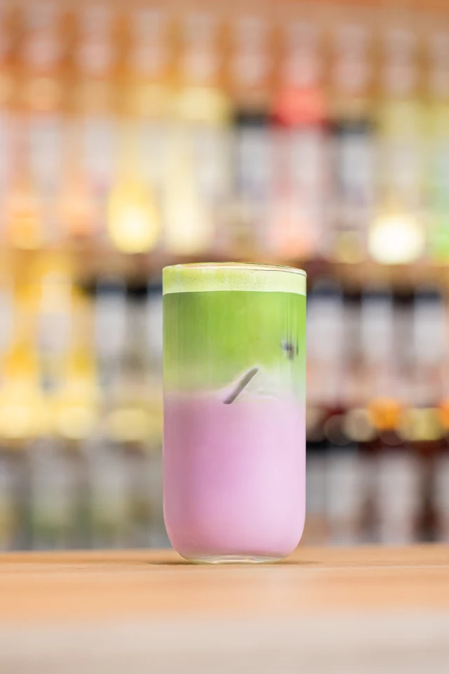 Studené vrstvené Ube matcha latte před barevnou barovou stěnou.